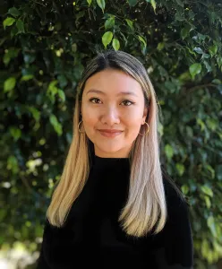 Bianca Lê with blonde hair and a black shirt standing in front of green trees.