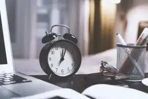 Black twin bell alarm clock on desk surrounded by laptop, books and pens.